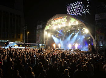 Illuminated stage with a band on it. In front of the stage there are hundreds of people standing and watching.